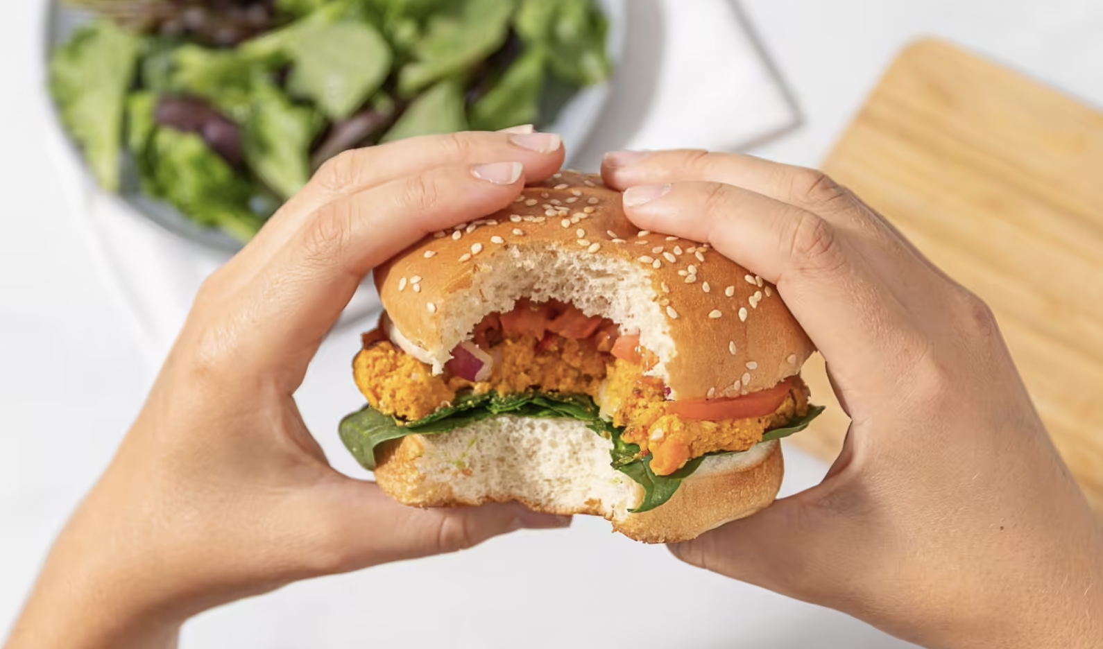 two hands holding a veggie burger with a bite out of it. salad can be seen in the background.